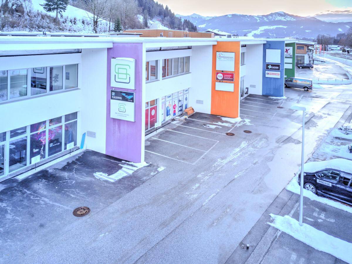 Geschäftsgebäude mit Parkplätzen im Winter, umgeben von schneebedeckten Bergen unter bewölktem Himmel.