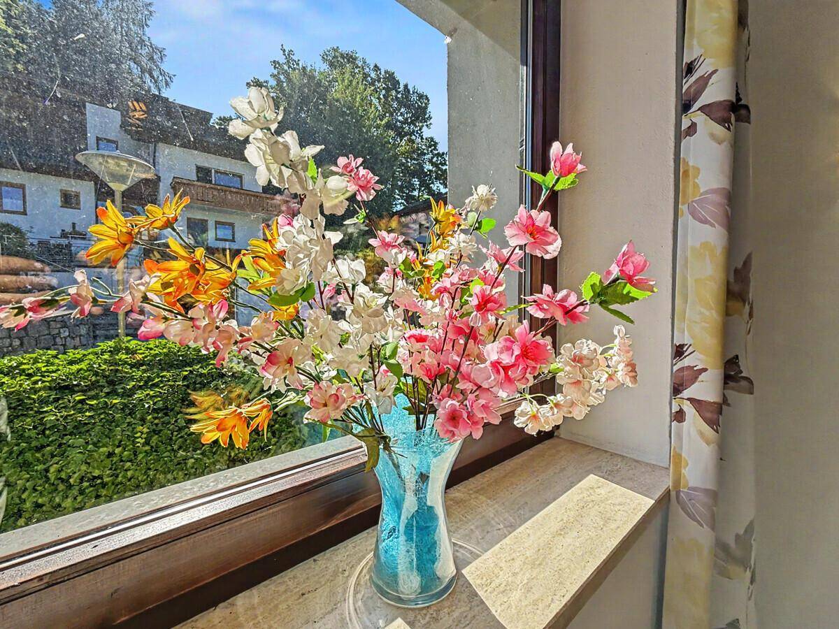 Fenster mit bunten Kunstblumen und Blick auf ein Nachbarhaus und grüne Umgebung.