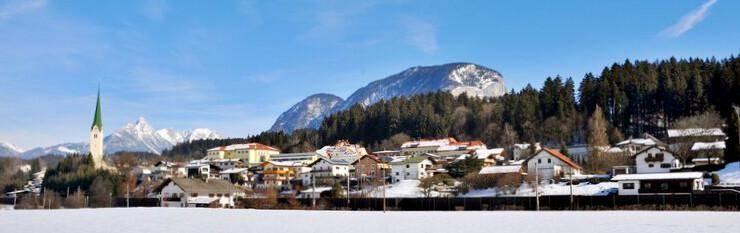 Panoramablick auf ein verschneites Dorf mit Kirchturm, umgeben von Bergen und Wäldern unter blauem Himmel.