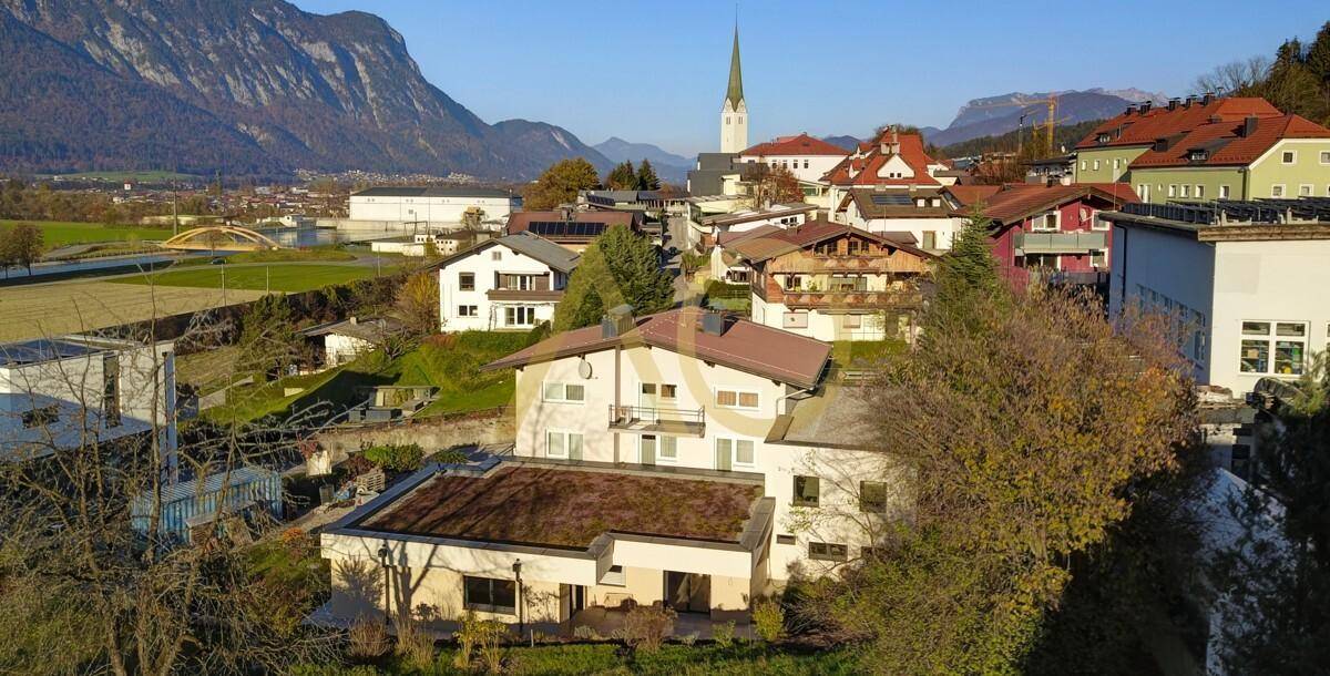 Luftaufnahme einer Wohnsiedlung mit Häusern, Bergen und einer Kirche im Hintergrund.