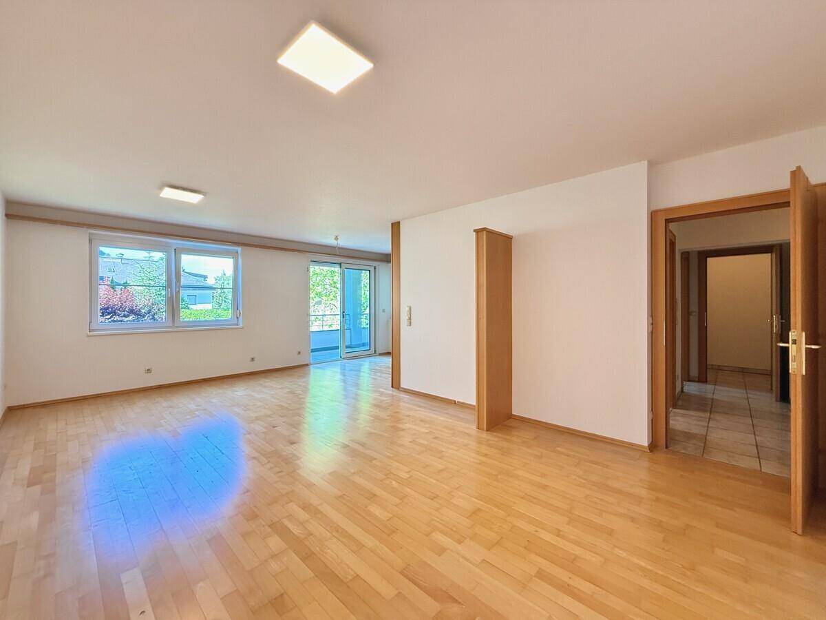 Helles, leeres Wohnzimmer mit Parkettboden und Zugang zu einem Balkon.