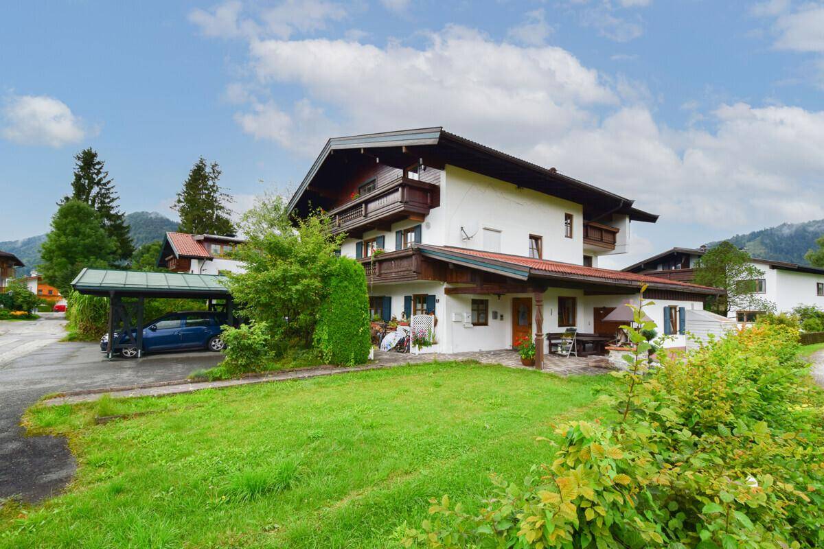 Traditionelles Haus im Alpenstil mit gepflegtem Garten, Carport und Bergblick unter blauem Himmel.