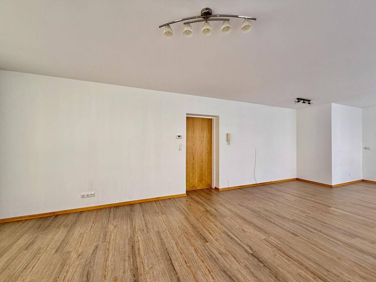 Leerer Wohnbereich mit hellem Laminatboden, weißen Wänden und einer Holztür.