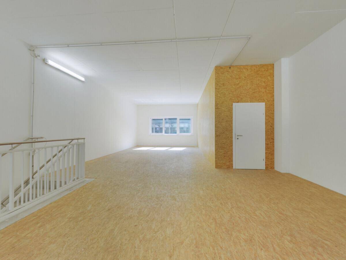 Leerer Gewerberaum mit OSB-Boden, weißen Wänden und einer Treppe, die nach unten führt, mit Fenster im Hintergrund.
