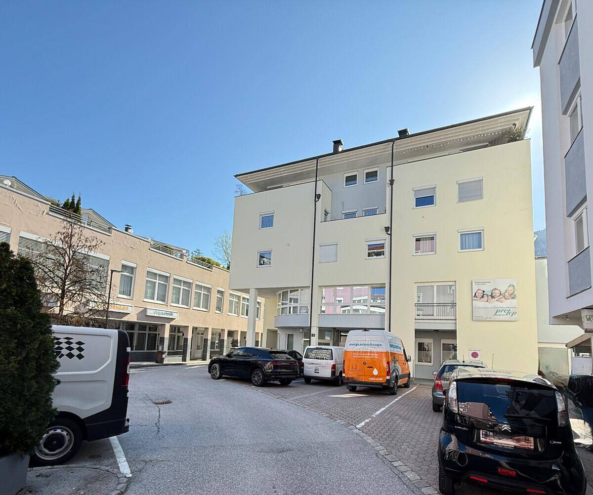 Modernes Gebäude mit heller Fassade und geparkten Fahrzeugen auf dem Vorplatz unter blauem Himmel.