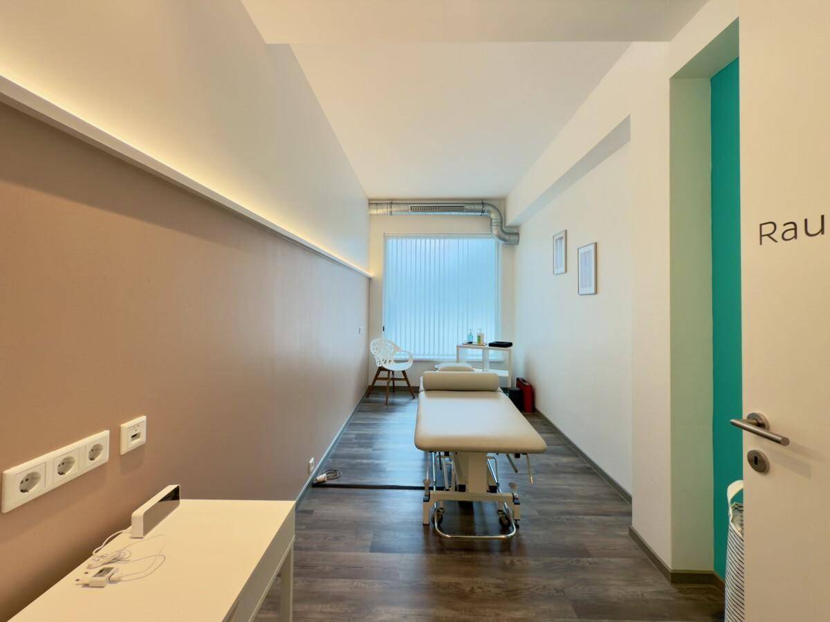 Heller Behandlungsraum mit Therapieliege, großem Fenster und indirekter Beleuchtung.