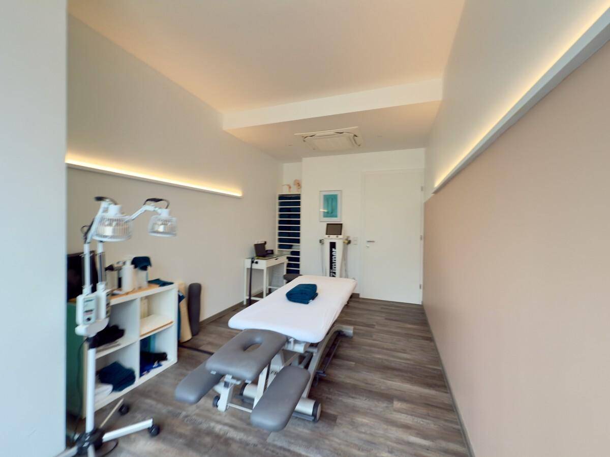 Langer Behandlungsraum mit Therapieliege, Regalen und indirekter Beleuchtung an den Wänden.