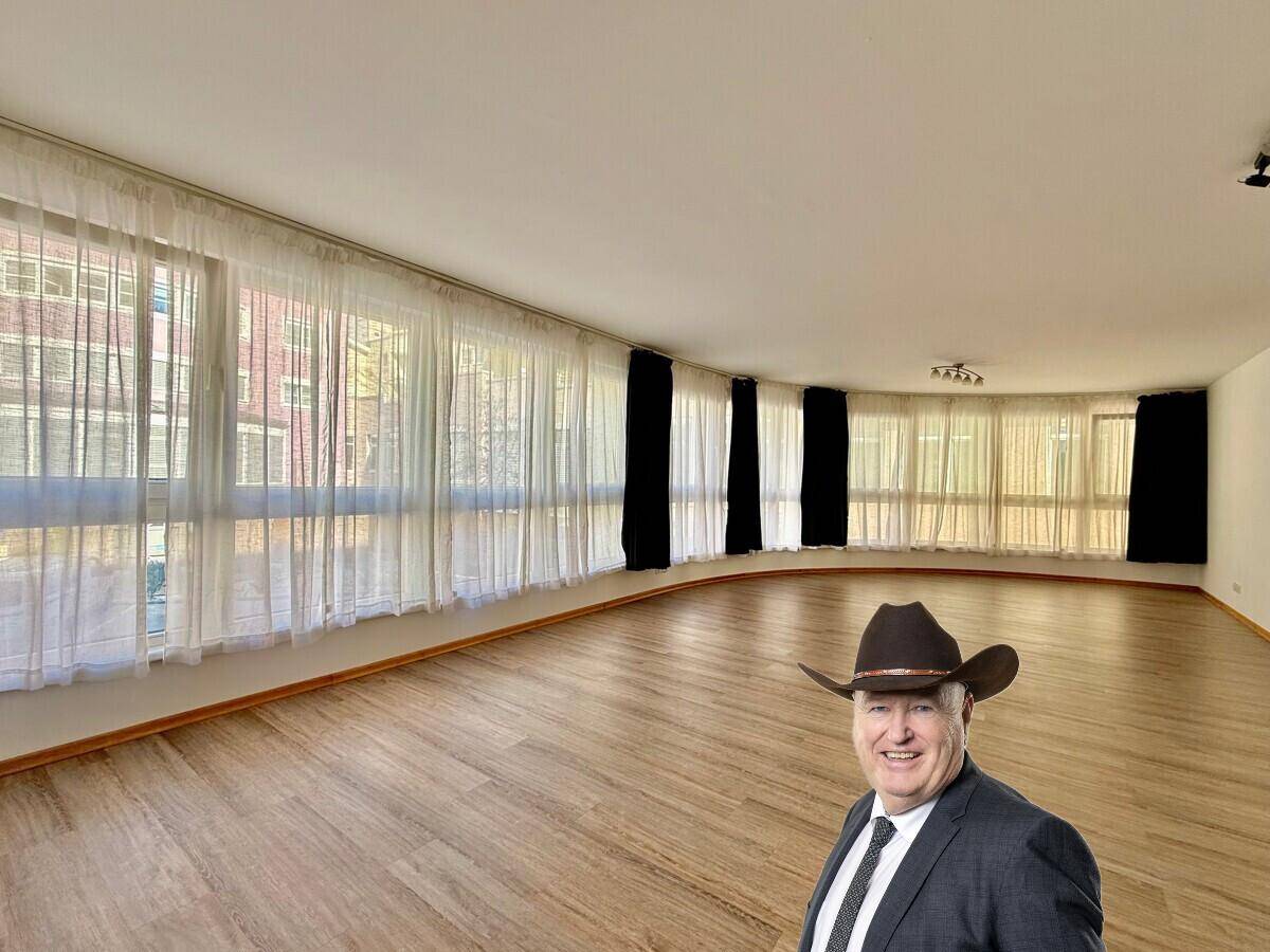 Geräumiges Wohnzimmer mit vielen Fenstern, hellem Laminatboden und einem Mann mit Hut im Vordergrund.