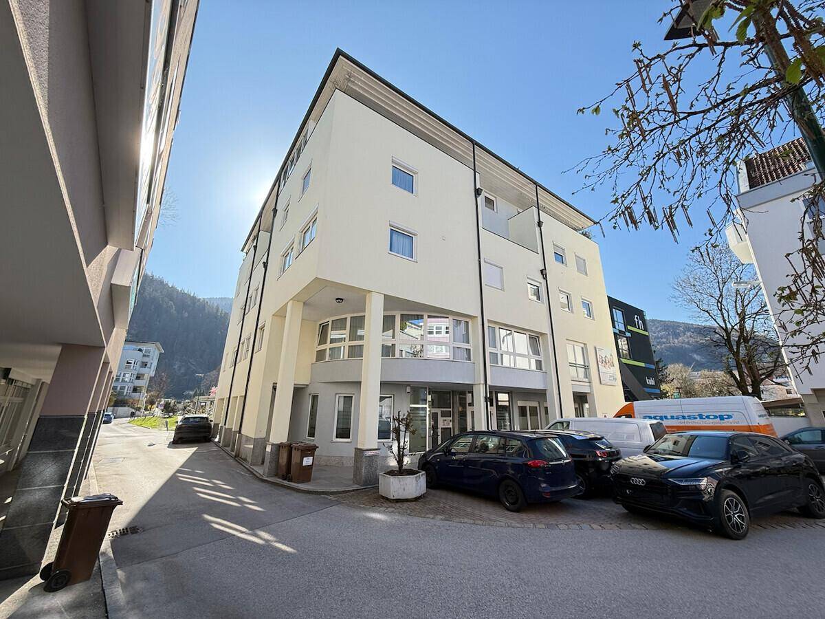 Modernes Gebäude mit heller Fassade, vielen Fenstern und geparkten Autos auf dem Vorplatz.