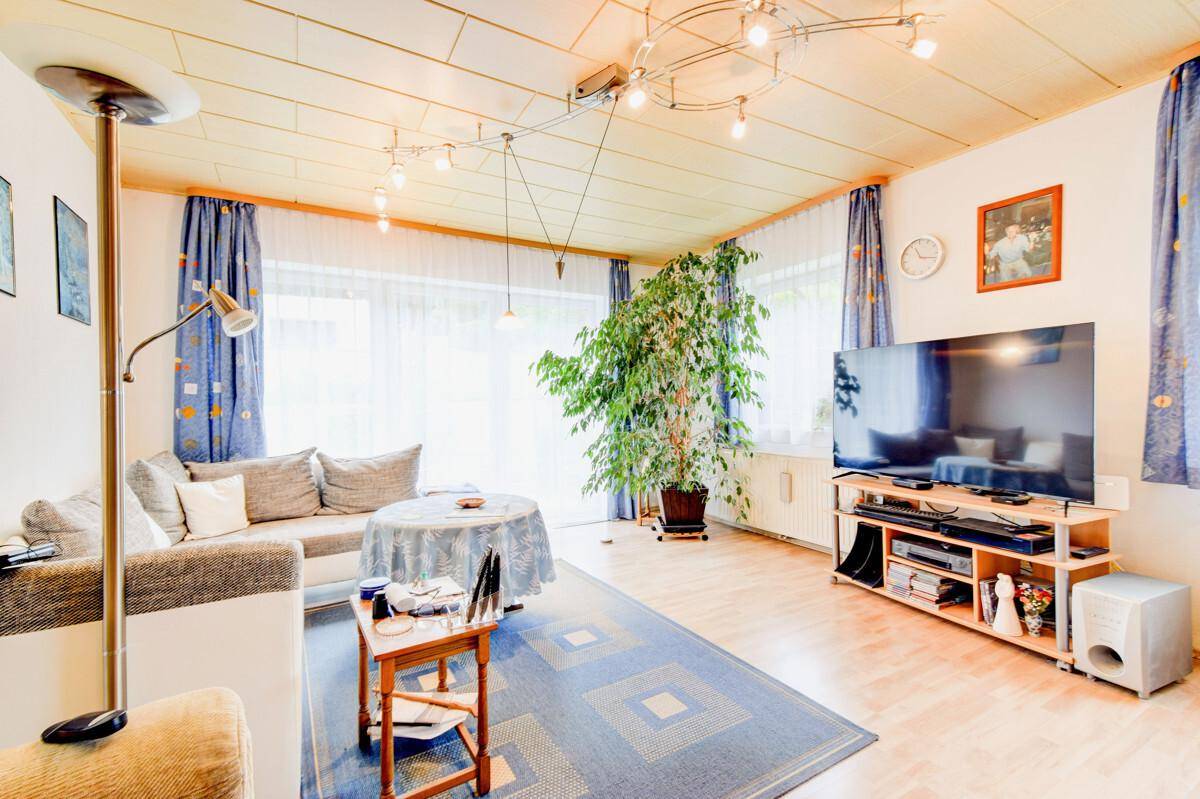Helles Wohnzimmer mit beigem Sofa, großem Fernseher und einer Zimmerpflanze, Holzbalkendecke.