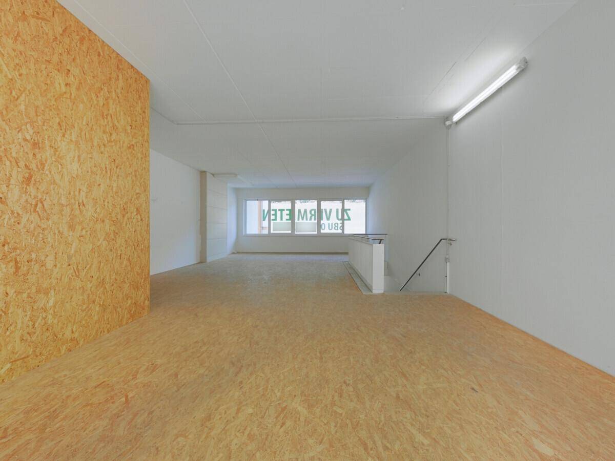 Geräumiger, leerer Gewerberaum mit OSB-Boden, weißen Wänden und großen Fenstern im Hintergrund, die Tageslicht hereinlassen.
