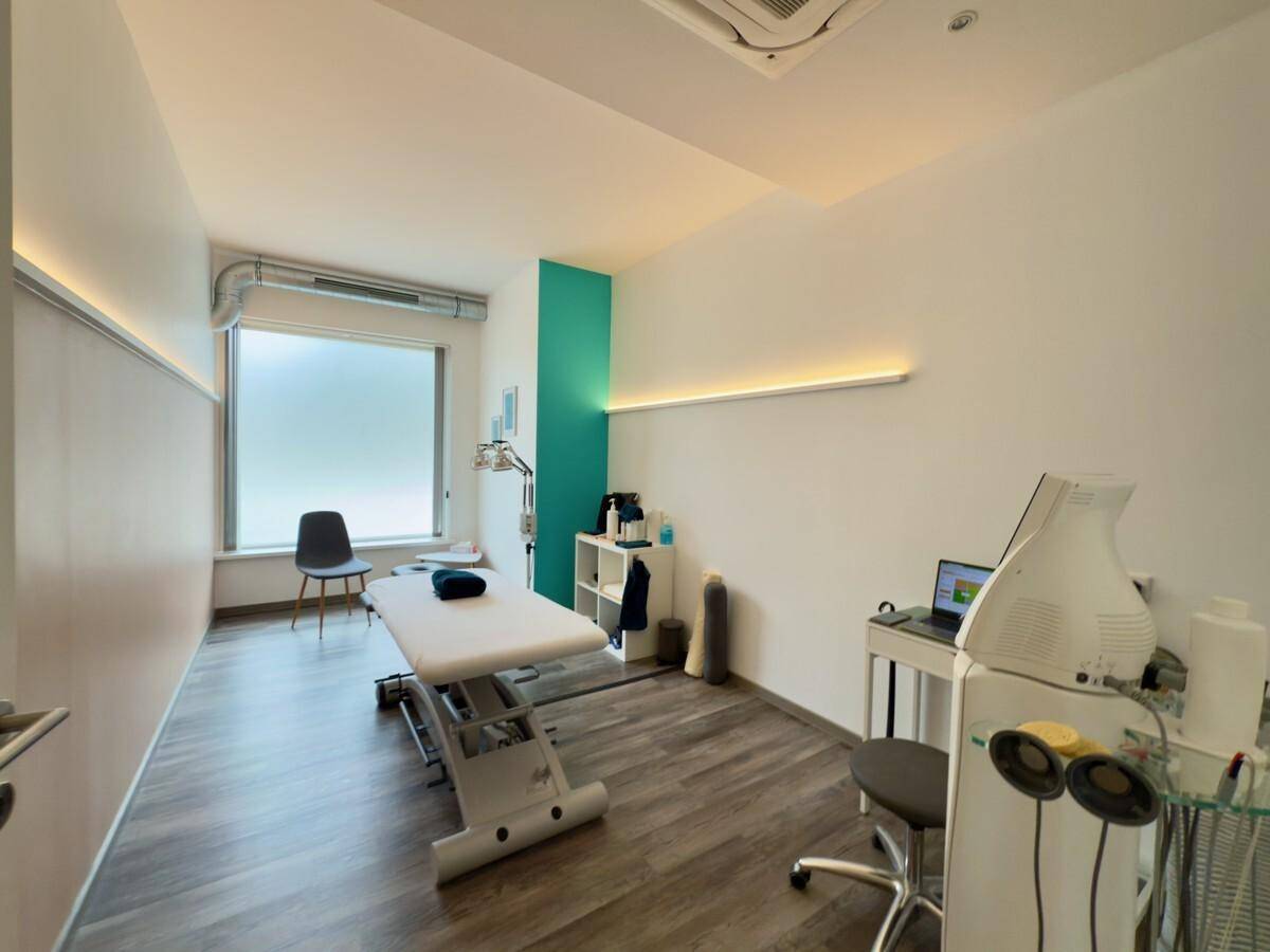 Behandlungsraum mit Therapieliege, großem Fenster und indirekter Beleuchtung an der Wand.