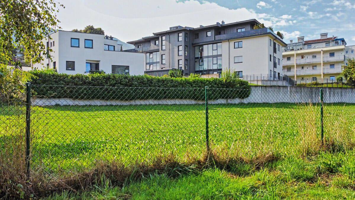 Offene Grünfläche mit Zaun und modernen Wohngebäuden unter blauem Himmel.