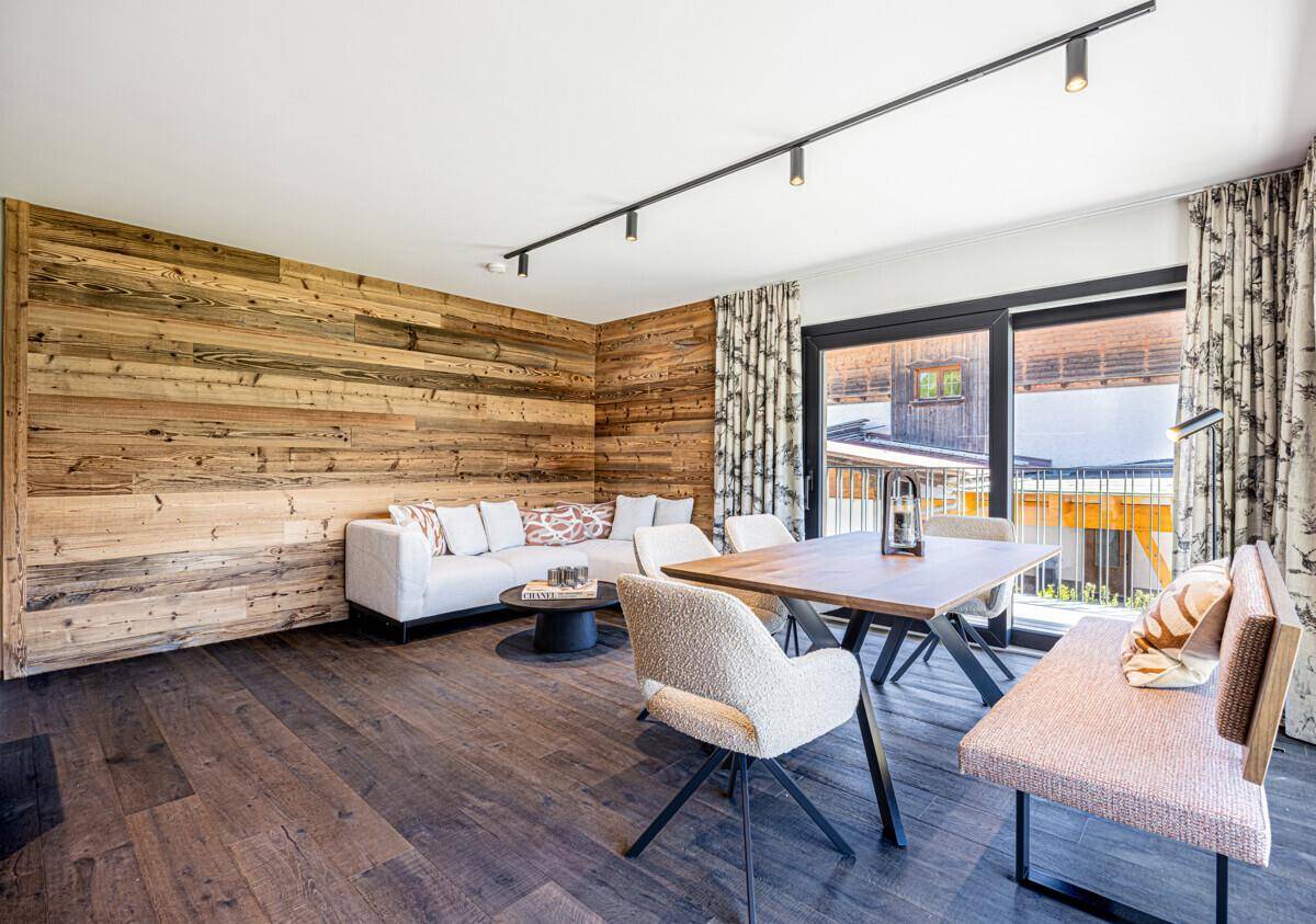 Gemütlicher Wohn-Essbereich mit Holzwand, bequemer Couch und Zugang zum Balkon durch große Fenster.