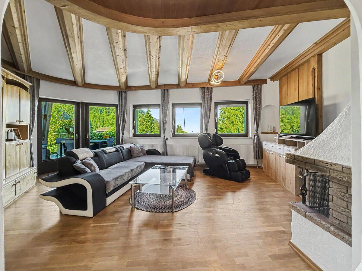 Geräumiges Wohnzimmer mit Holzbalkendecke, Kamin und großen Fenstern mit Blick ins Grüne.