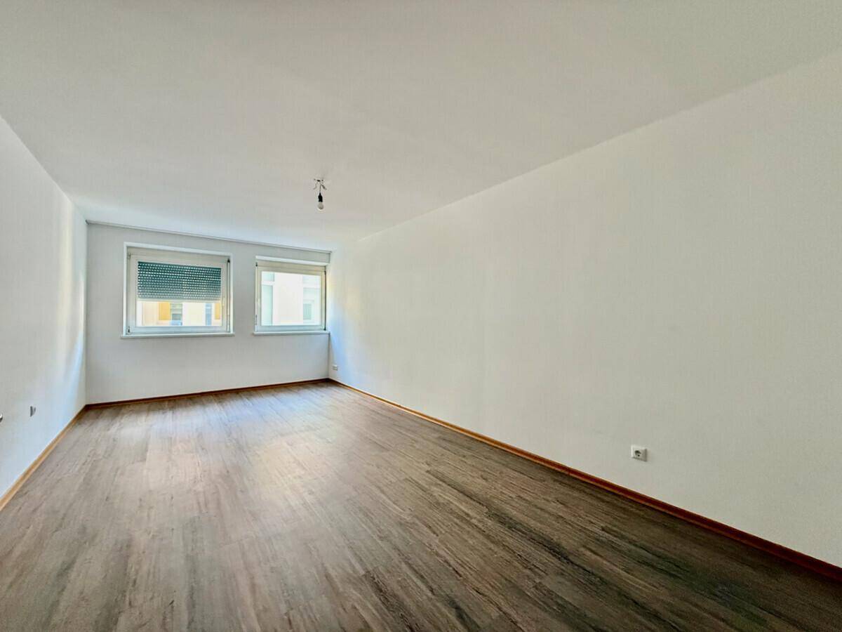 Leeres Zimmer mit Laminatboden, weißen Wänden und zwei Fenstern mit Rollläden.