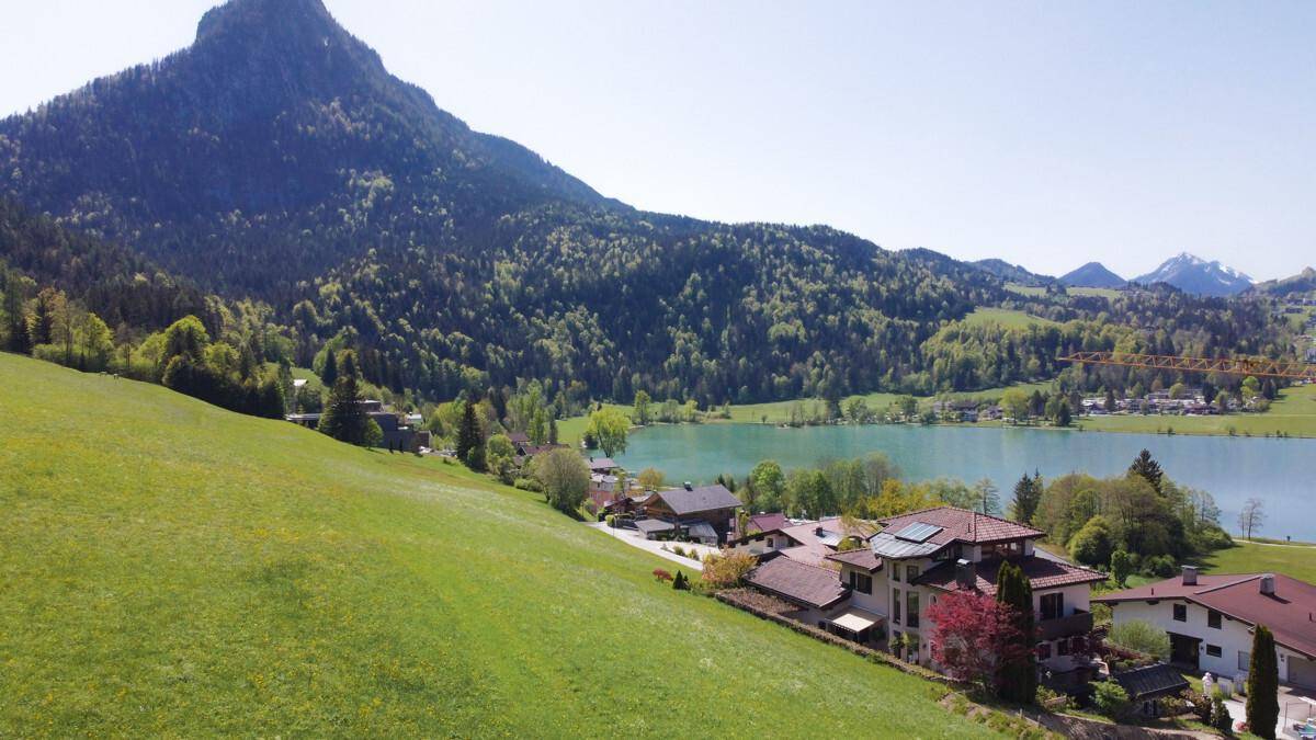 Panoramablick auf einen idyllischen See, umgeben von grünen Hügeln und majestätischen Bergen. Mehrere Häuser sind harmonisch in die Landschaft eingebettet.
