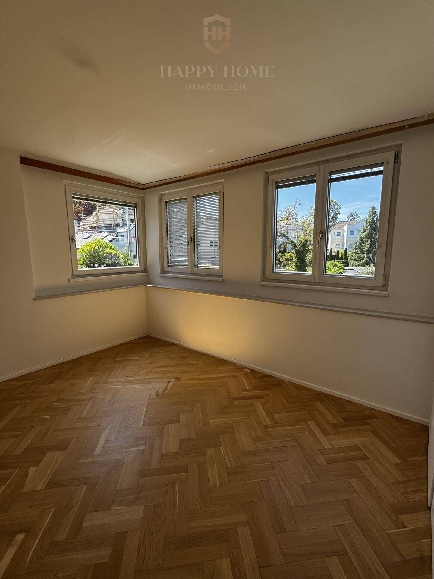 Leeres Zimmer mit Fischgrätparkett und mehreren Fenstern, die viel Tageslicht hereinlassen.