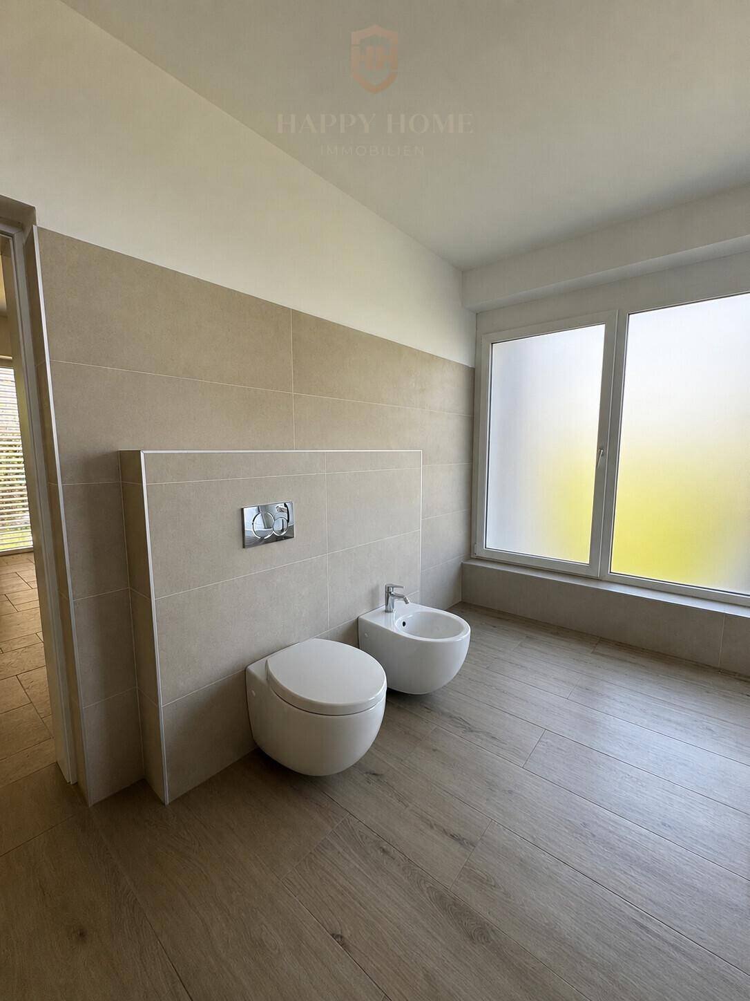 Modernes Badezimmer mit Wand-WC, Bidet und großen, undurchsichtigen Fenstern für Privatsphäre.