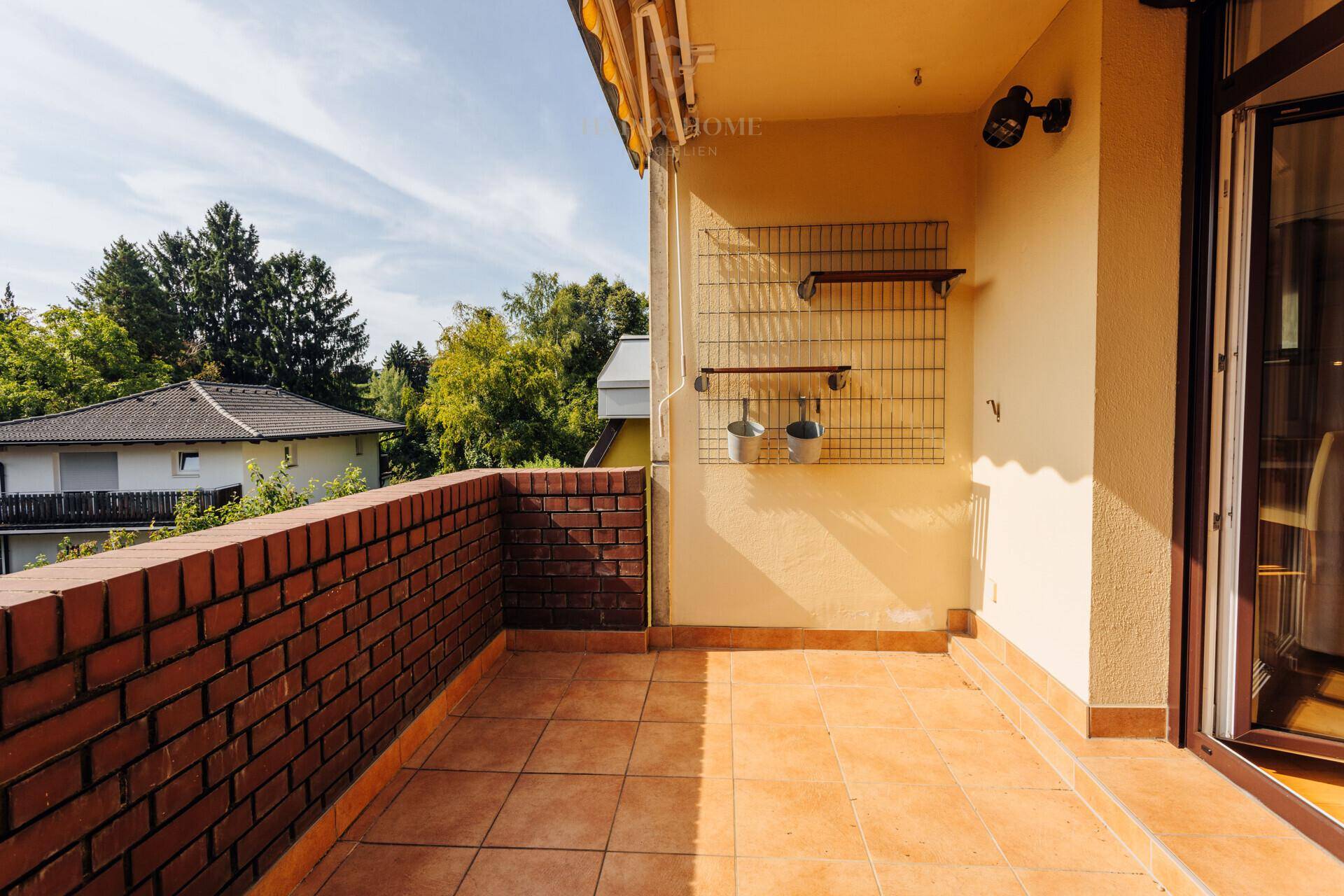 Sonniger Balkon mit Ziegelmauer, Fliesenboden und Blick ins Grüne.