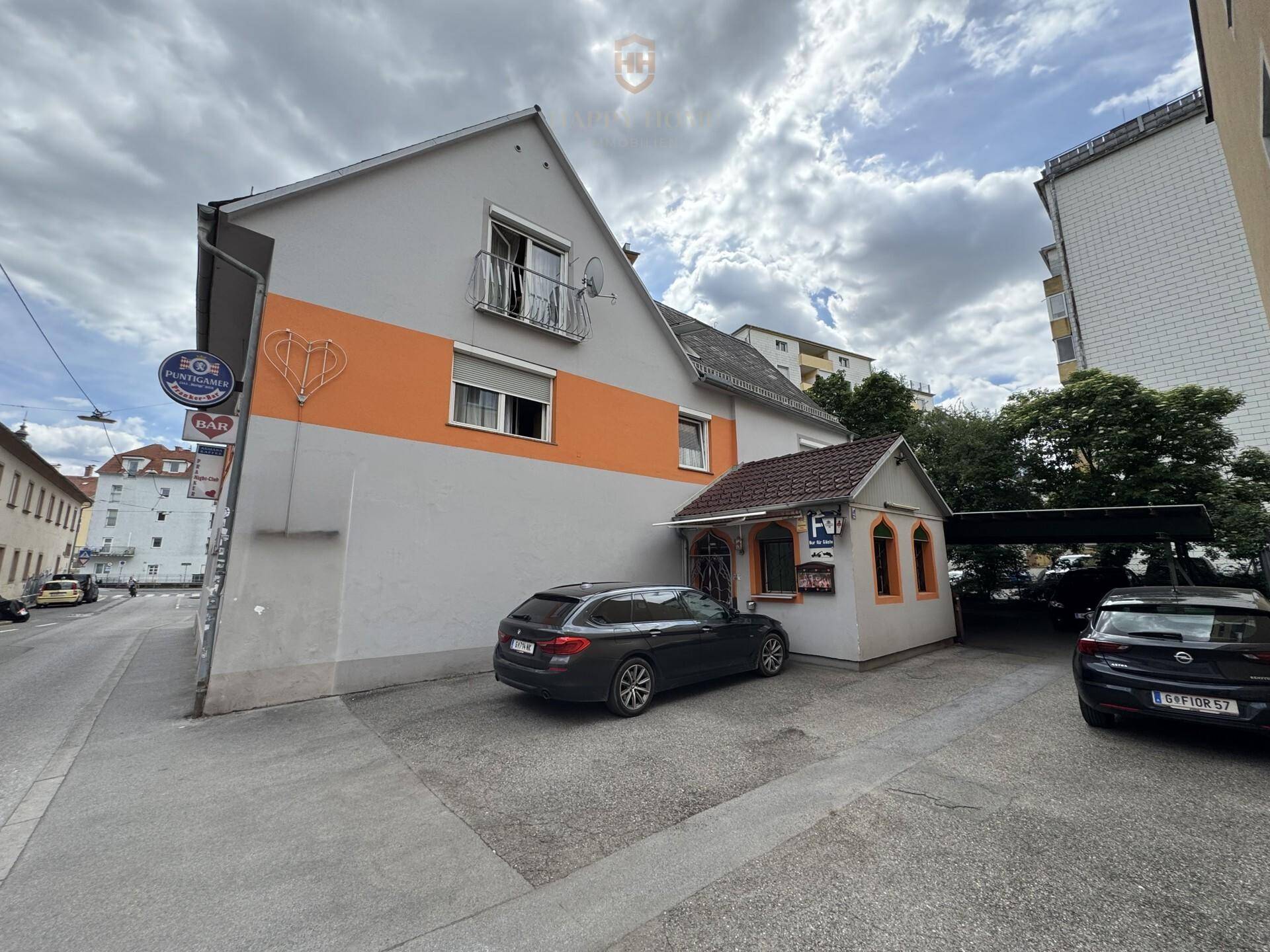 Fassade des Gebäudes mit Parkplätzen und einem kleinen Anbau an einer städtischen Straße.