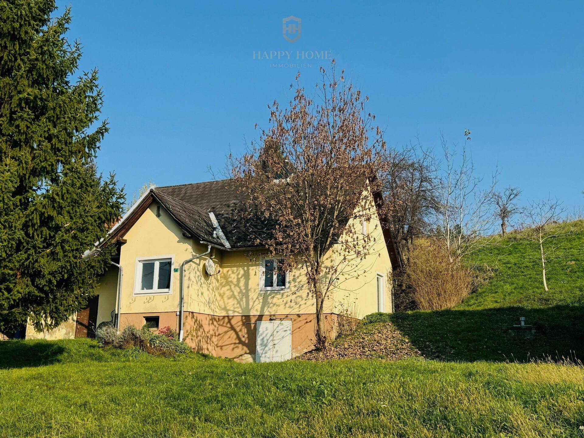 Gelbes Landhaus mit Satteldach und Fenstern, umgeben von grüner Wiese und Bäumen.