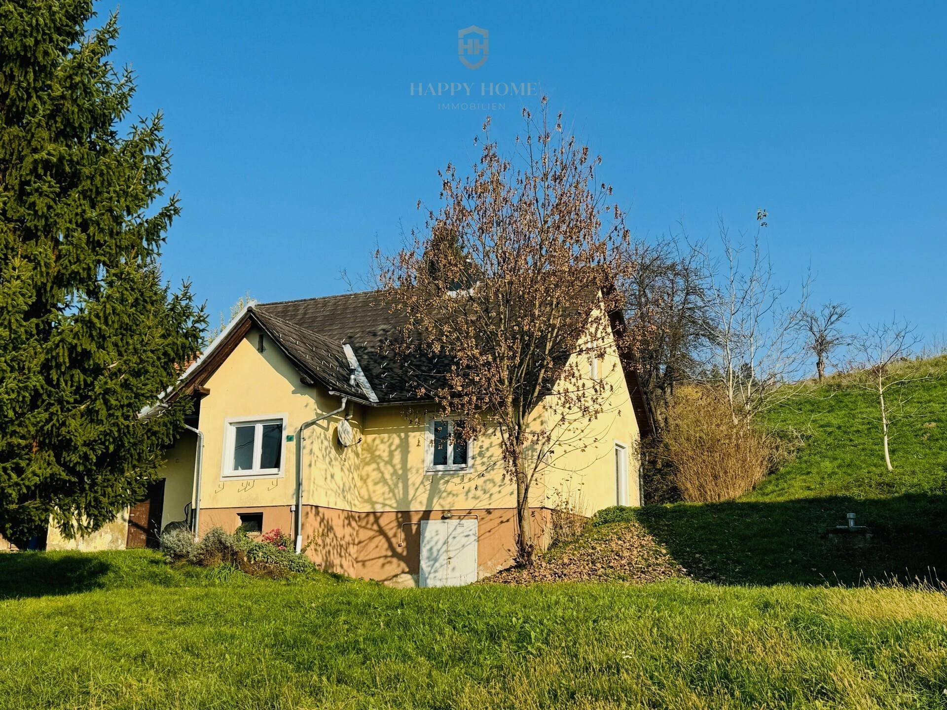 Gelbes Landhaus mit Satteldach und Fenstern, umgeben von grüner Wiese und Bäumen.