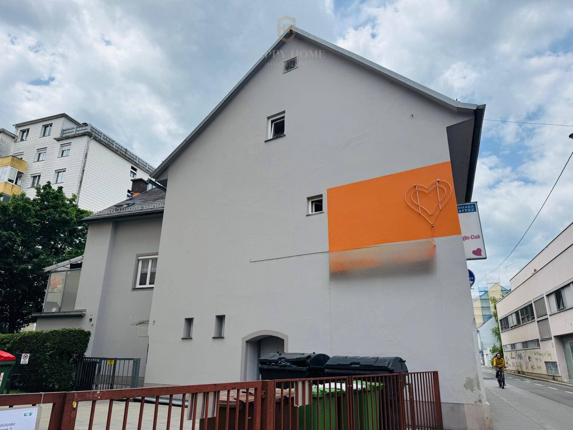 Seitenansicht des Gebäudes mit orangefarbenem Akzent und Mülltonnenbereich an der Straße.