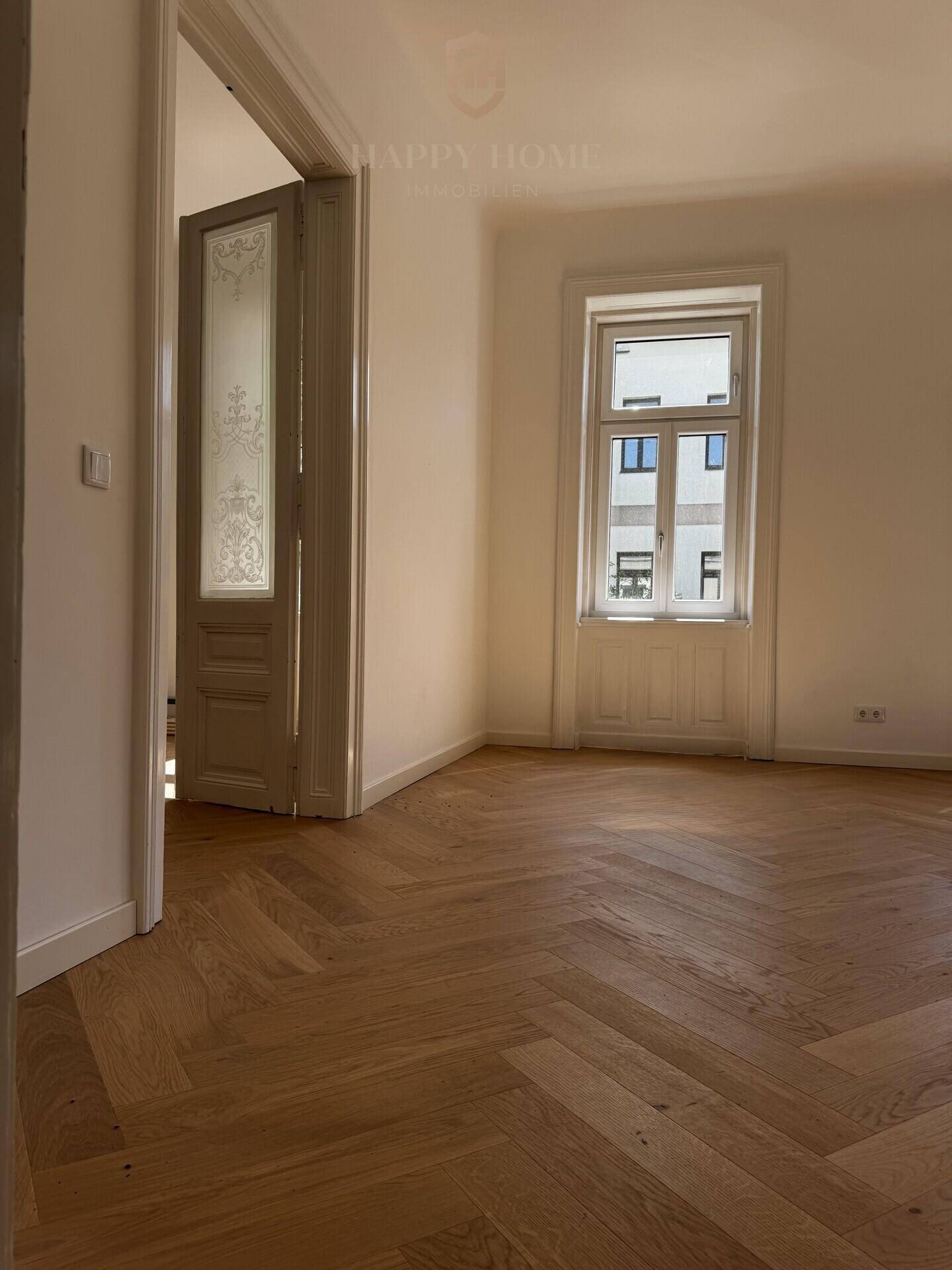 Geräumiges Zimmer mit Fischgrätparkett und hohen Fenstern, die auf die Straße blicken.