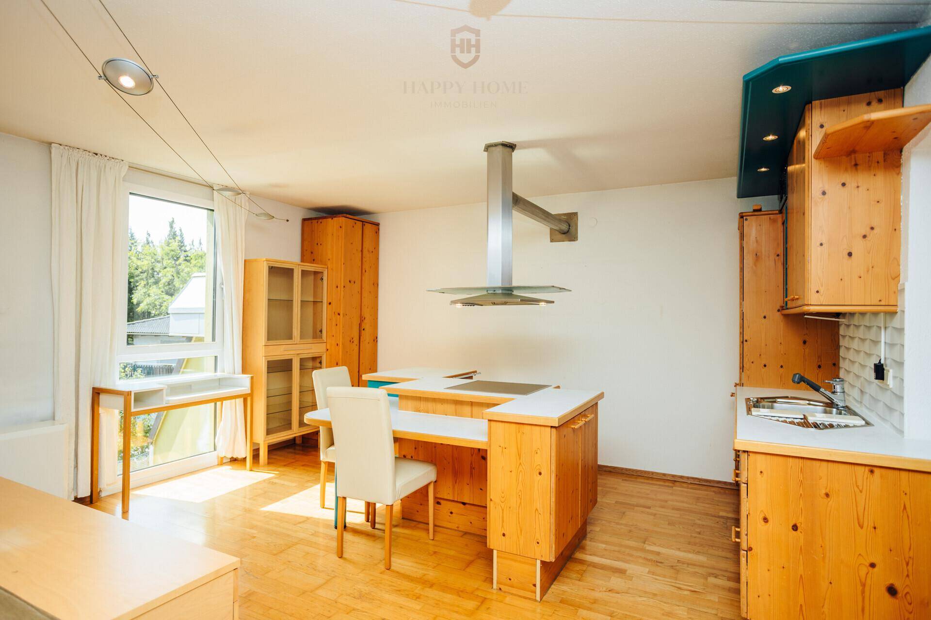 Helle Küche mit Kochinsel, Essbereich und Blick ins Grüne durch große Fenster.