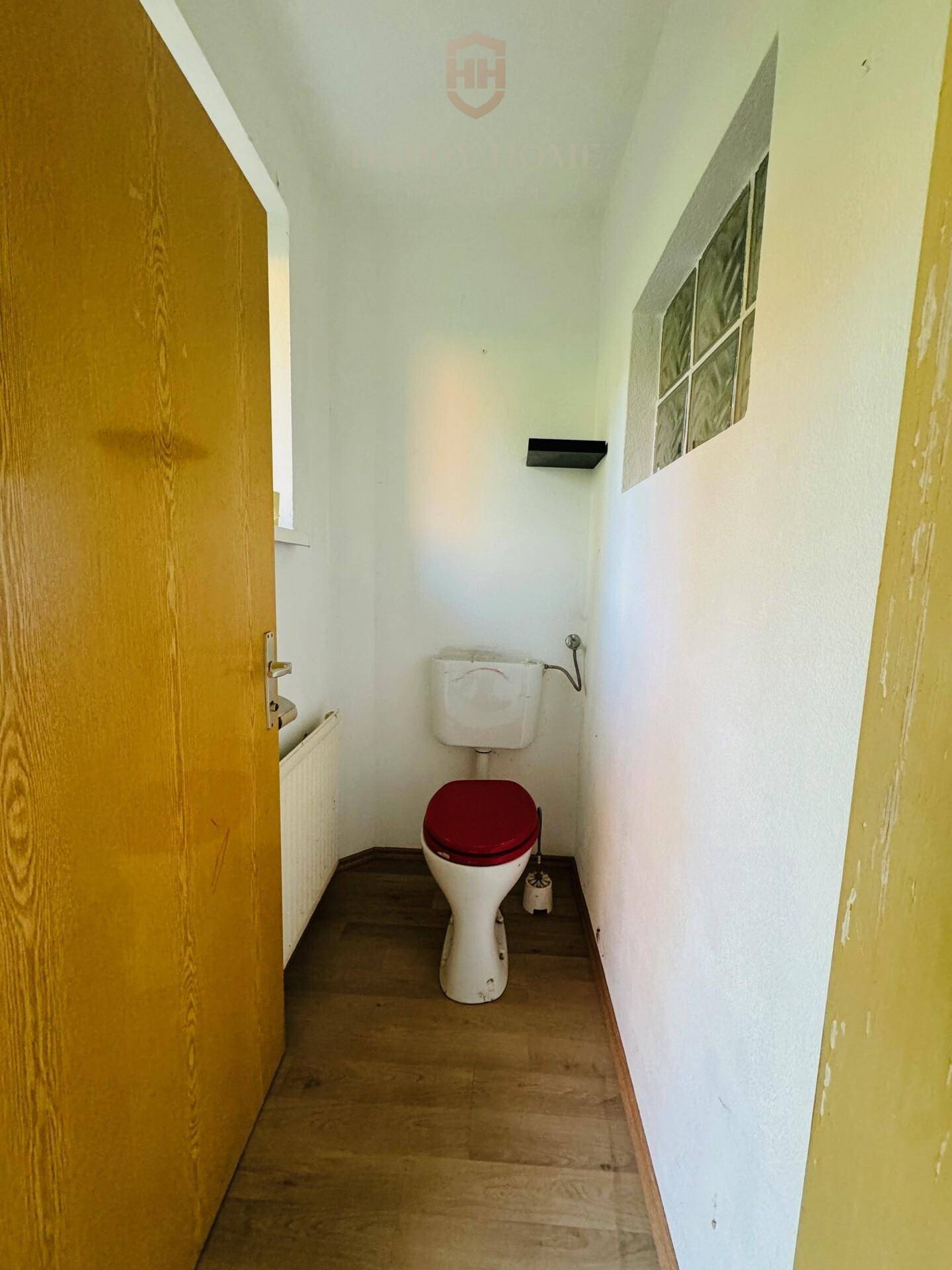Einfache Gästetoilette mit Fenster und rotem Toilettensitz, funktional und hell.