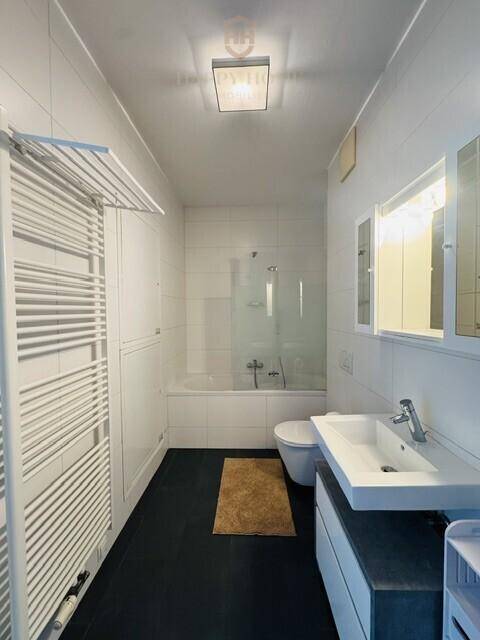 Helles Badezimmer mit weißen Fliesen, Badewanne mit Duschwand und beheiztem Handtuchhalter.