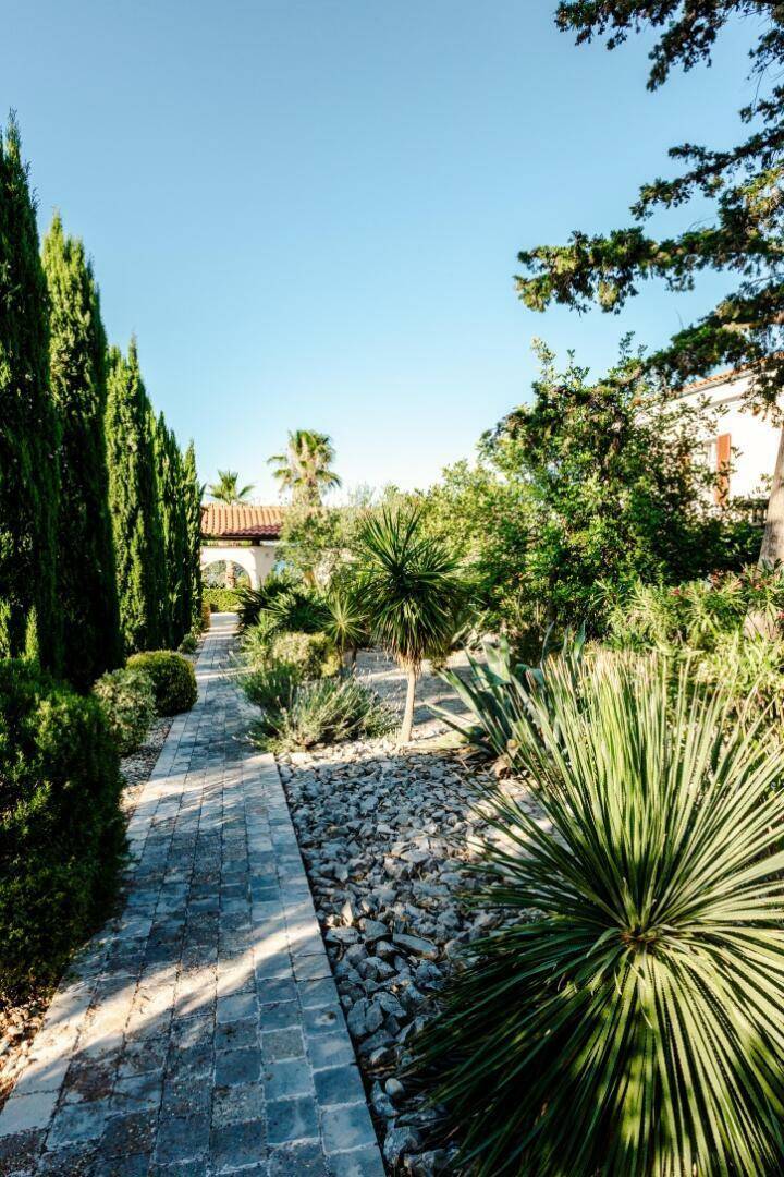 Sonniger Gartenweg mit Zypressen und mediterranen Pflanzen, der zu einem Torbogen führt.
