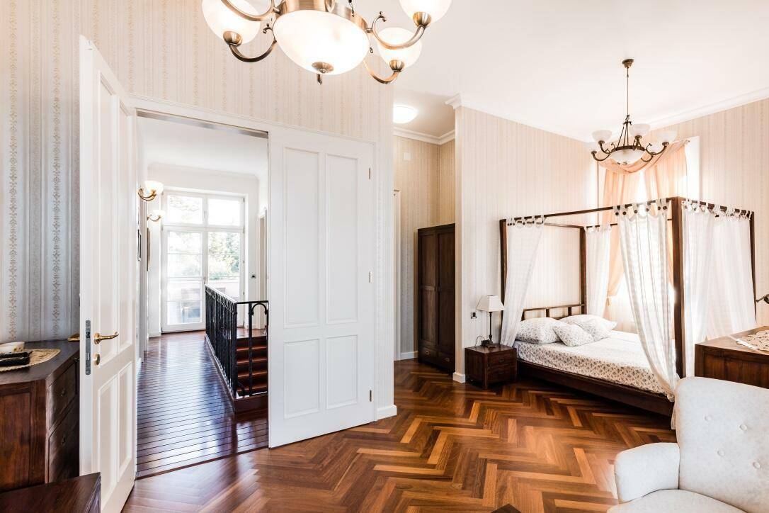 Helles Schlafzimmer mit Himmelbett, Fischgrätparkett und Zugang zu einem Flur mit Balkon.