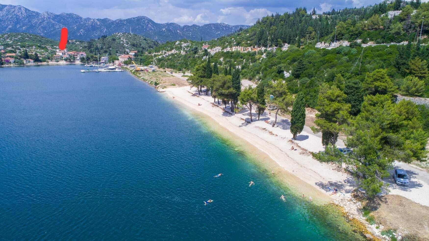 Panoramablick auf eine malerische Bucht mit Sandstrand, klarem Meer und bewaldeten Hügeln im Hintergrund.