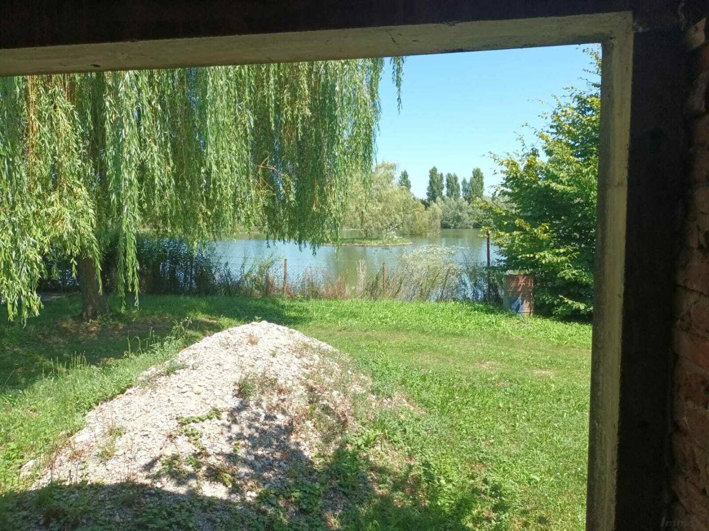 Blick aus einem unfertigen Innenraum auf einen See mit üppiger Vegetation und einem Haufen Kies.