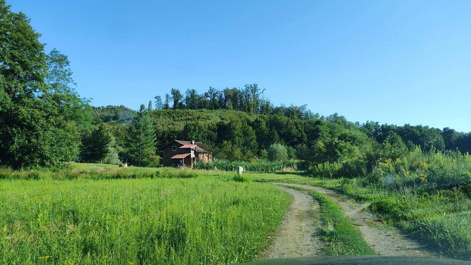 Landhaus in grüner Umgebung mit Feldweg und bewaldeten Hügeln im Hintergrund.