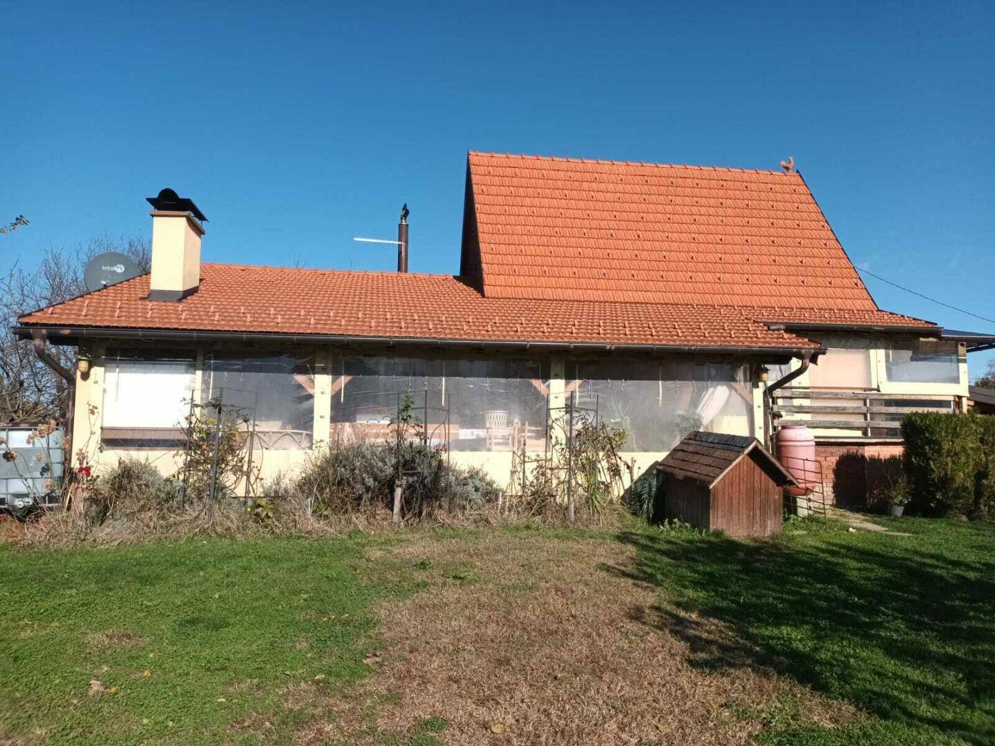 Hausfassade mit Ziegeldach, verglastem Anbau und grünem Vorgarten.