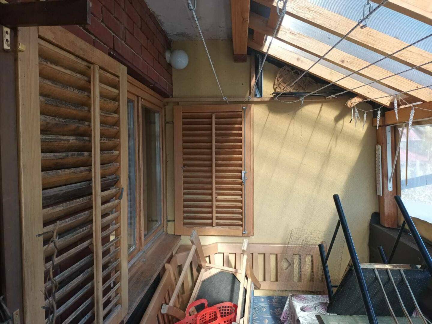 Überdachter Bereich mit Holzfensterläden und transparentem Dach, genutzt als Abstellfläche.