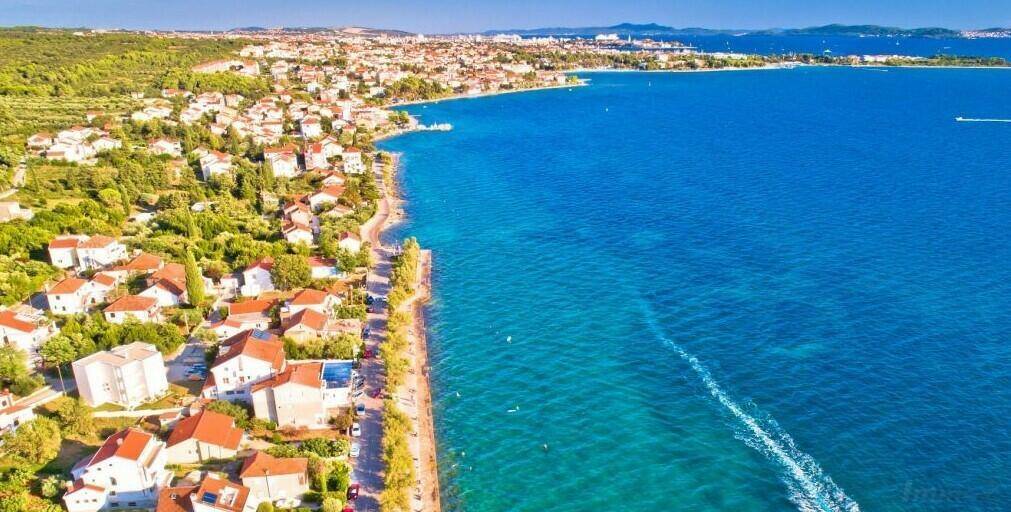 Weitläufige Luftaufnahme einer Küstenlinie mit Stadtbebauung und blauem Meer unter strahlendem Himmel.