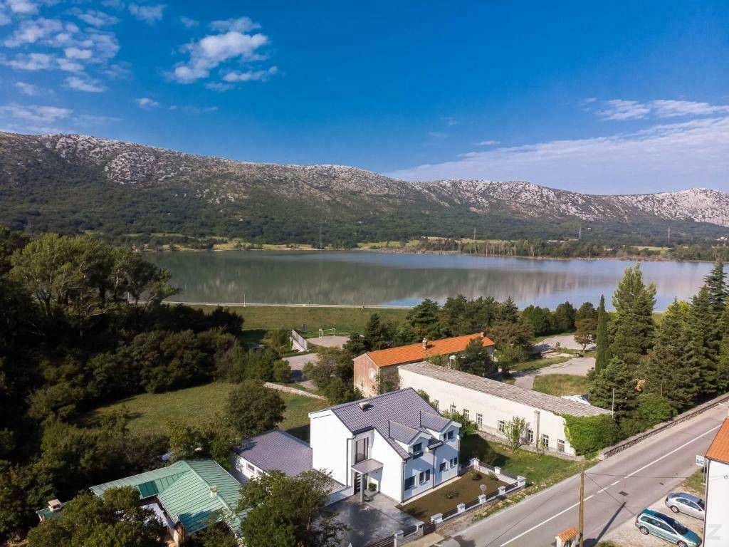 Luftaufnahme eines modernen Hauses in der Nähe eines Sees, umgeben von Bergen und grüner Landschaft.