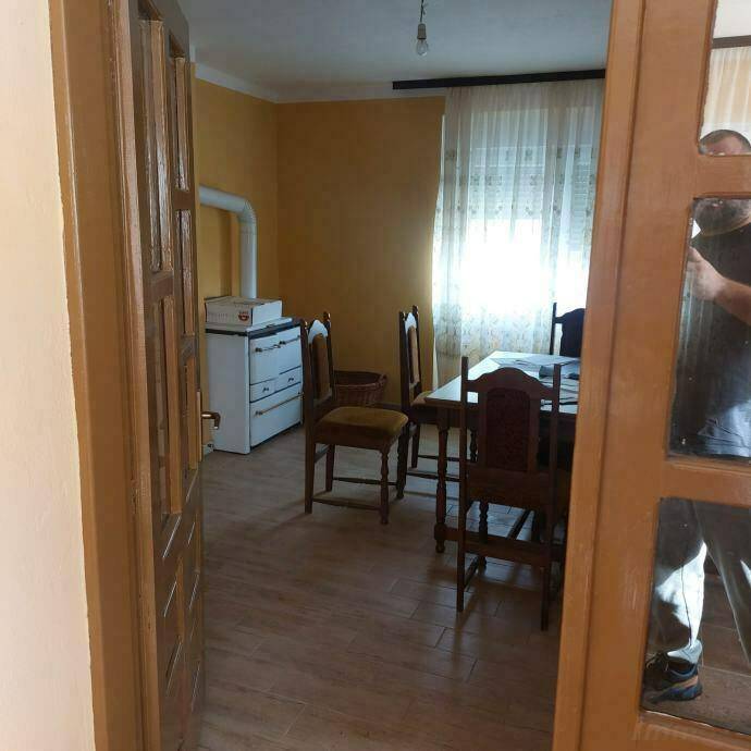 Esszimmer mit Holztisch, Stühlen und einem traditionellen Ofen, hell durch Fenster.