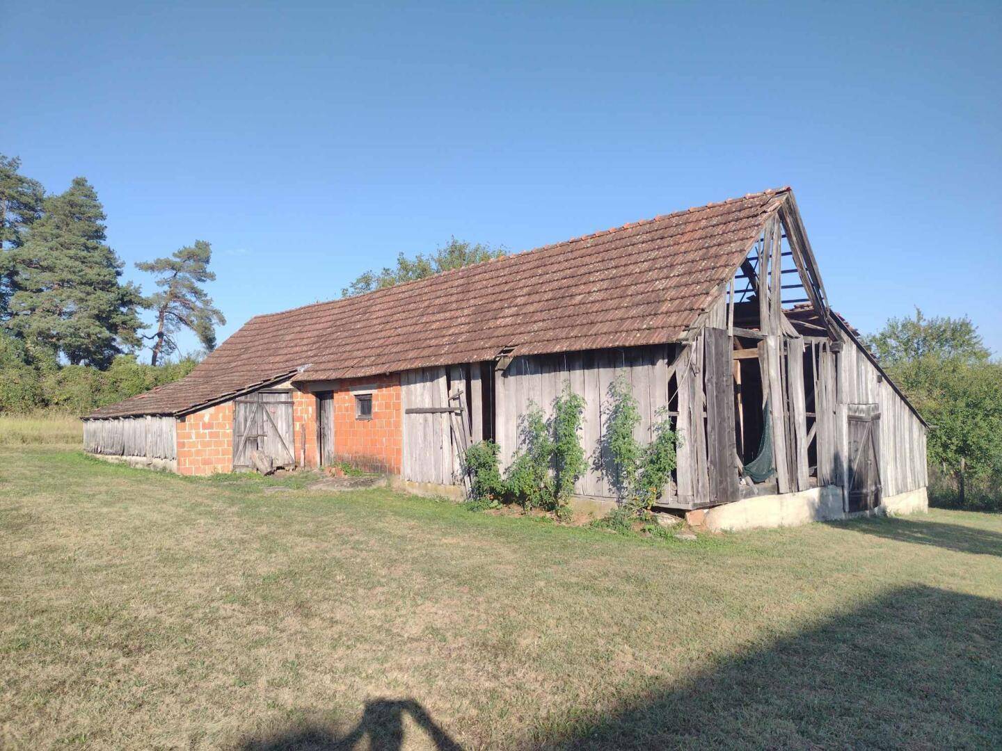Alte Holzscheune mit Ziegeldach und teilweise verfallener Fassade in ländlicher Umgebung.
