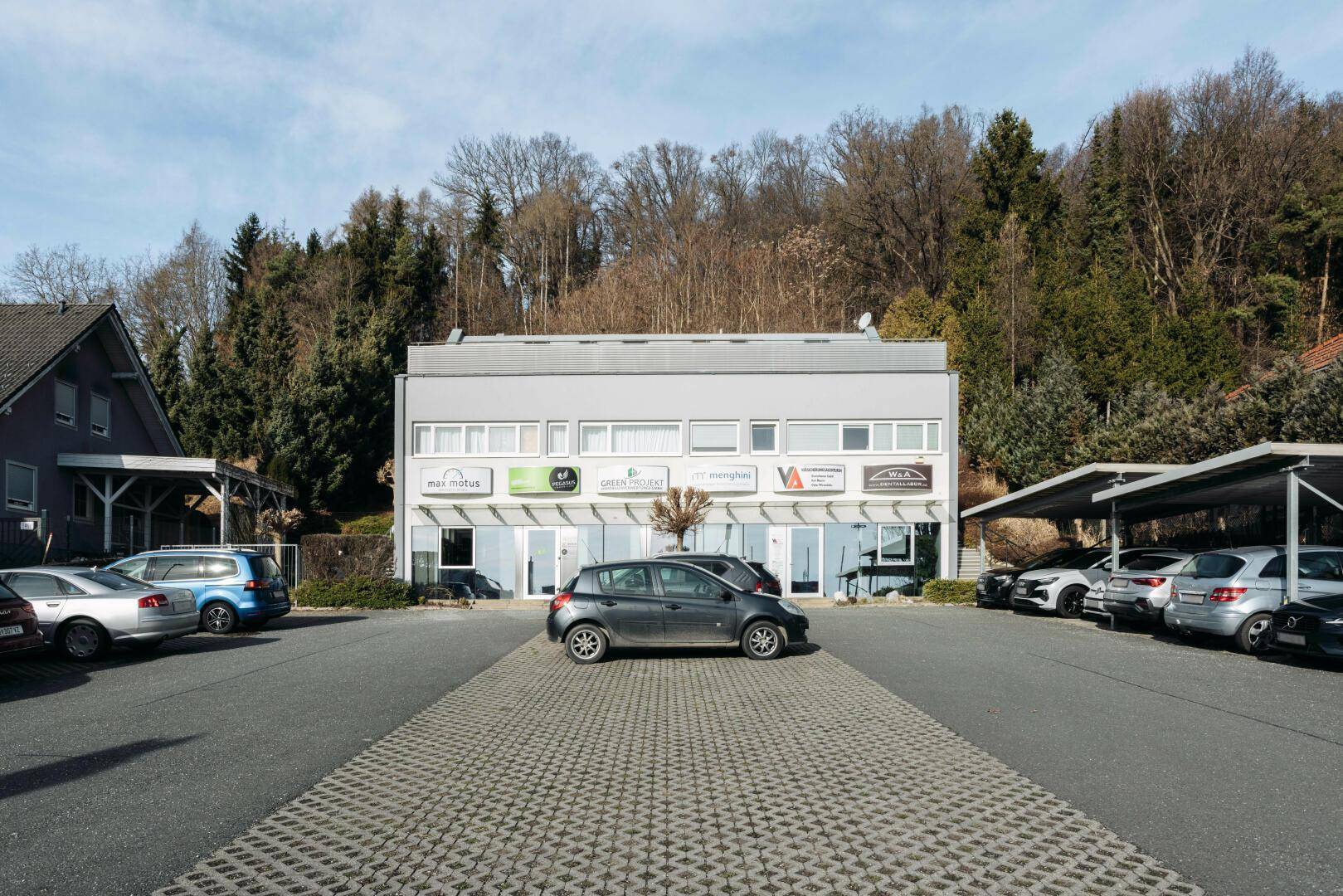 Geschäftsgebäude mit Parkplätzen und Carports vor einer bewaldeten Hügellandschaft.