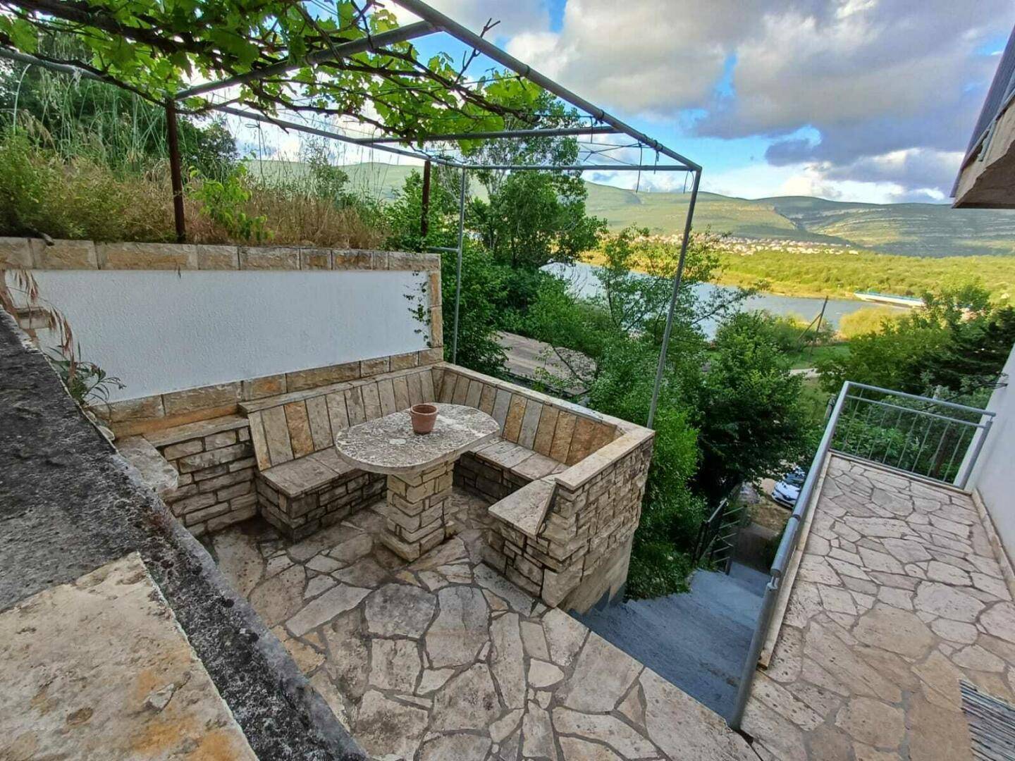 Gemütlicher Sitzbereich im Freien aus Stein mit Pergola und weitem Blick auf den See und die Berge.