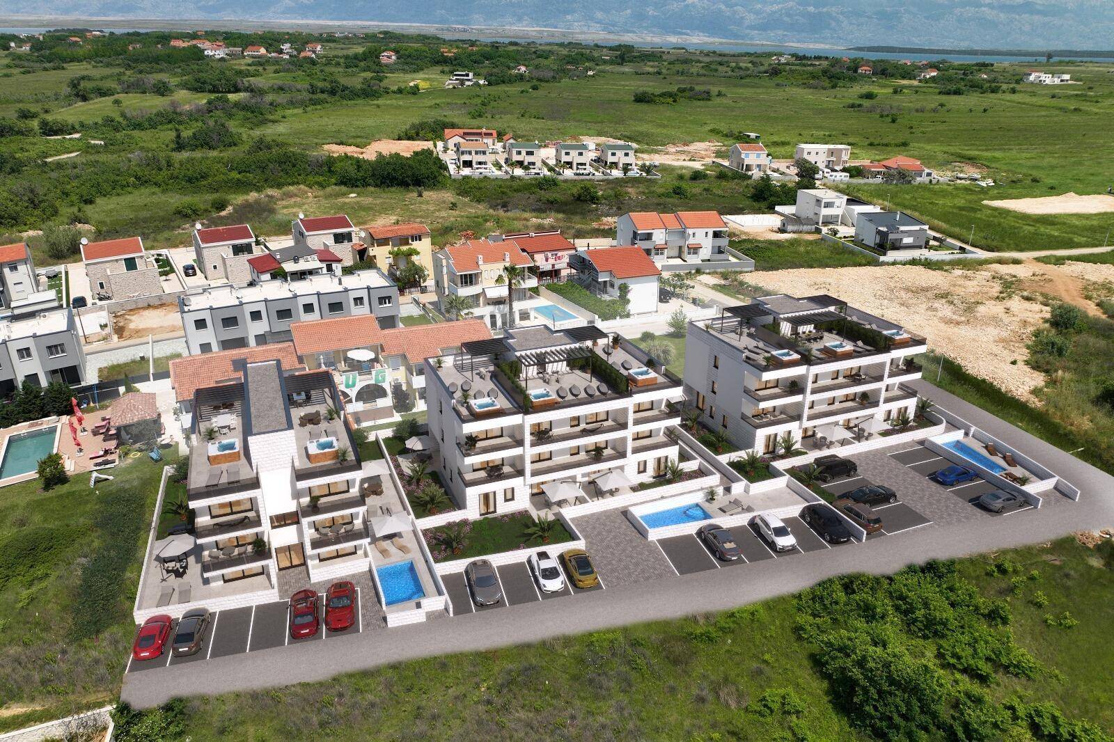 Luftaufnahme eines modernen Wohnkomplexes mit mehreren Gebäuden, Pools und Parkplätzen, umgeben von grüner Landschaft und Meerblick.