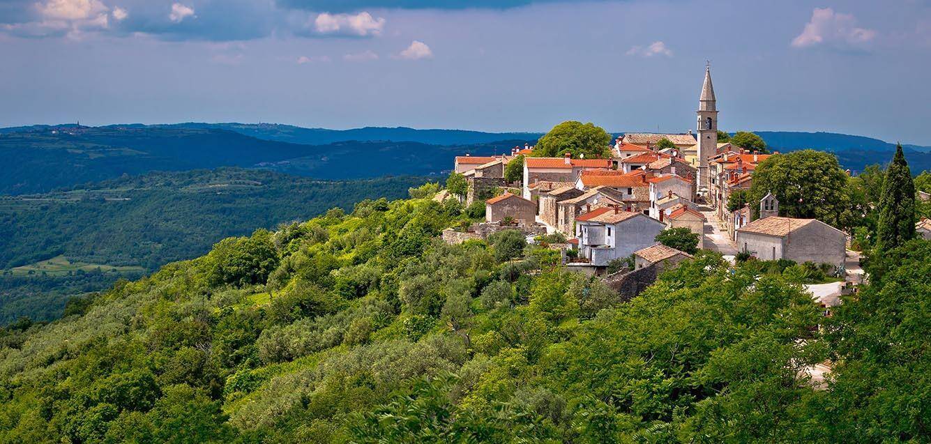 Historisches Dorf auf einem Hügel, umgeben von üppiger grüner Landschaft unter blauem Himmel.