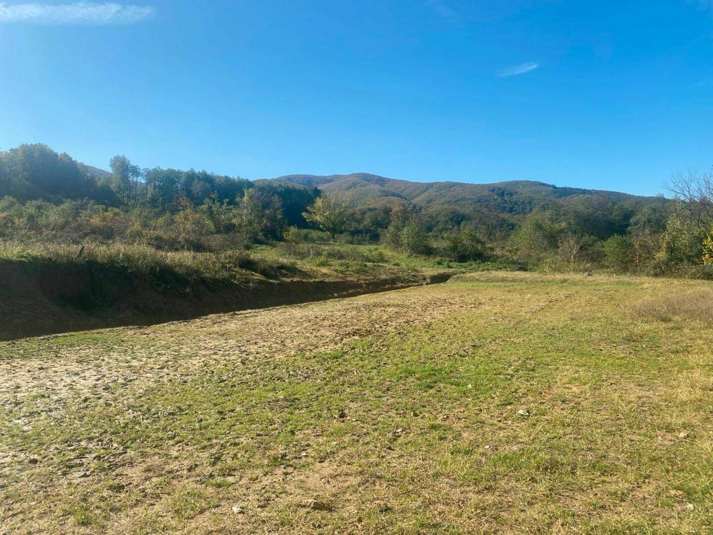 Weitläufiges Grundstück mit Gras und Erde, umgeben von bewaldeten Hügeln unter blauem Himmel.