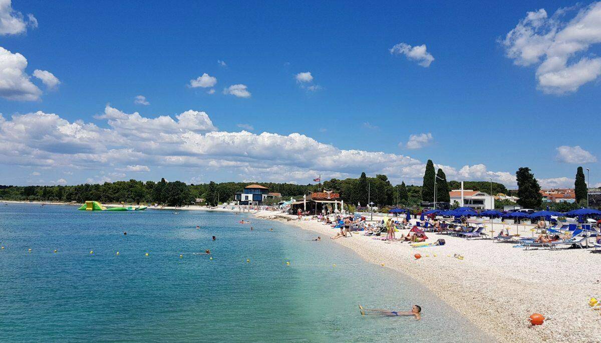 Belebter Sandstrand mit vielen Sonnenanbetern, bunten Sonnenschirmen und kristallklarem, blauem Meerwasser an einem sonnigen Tag.
