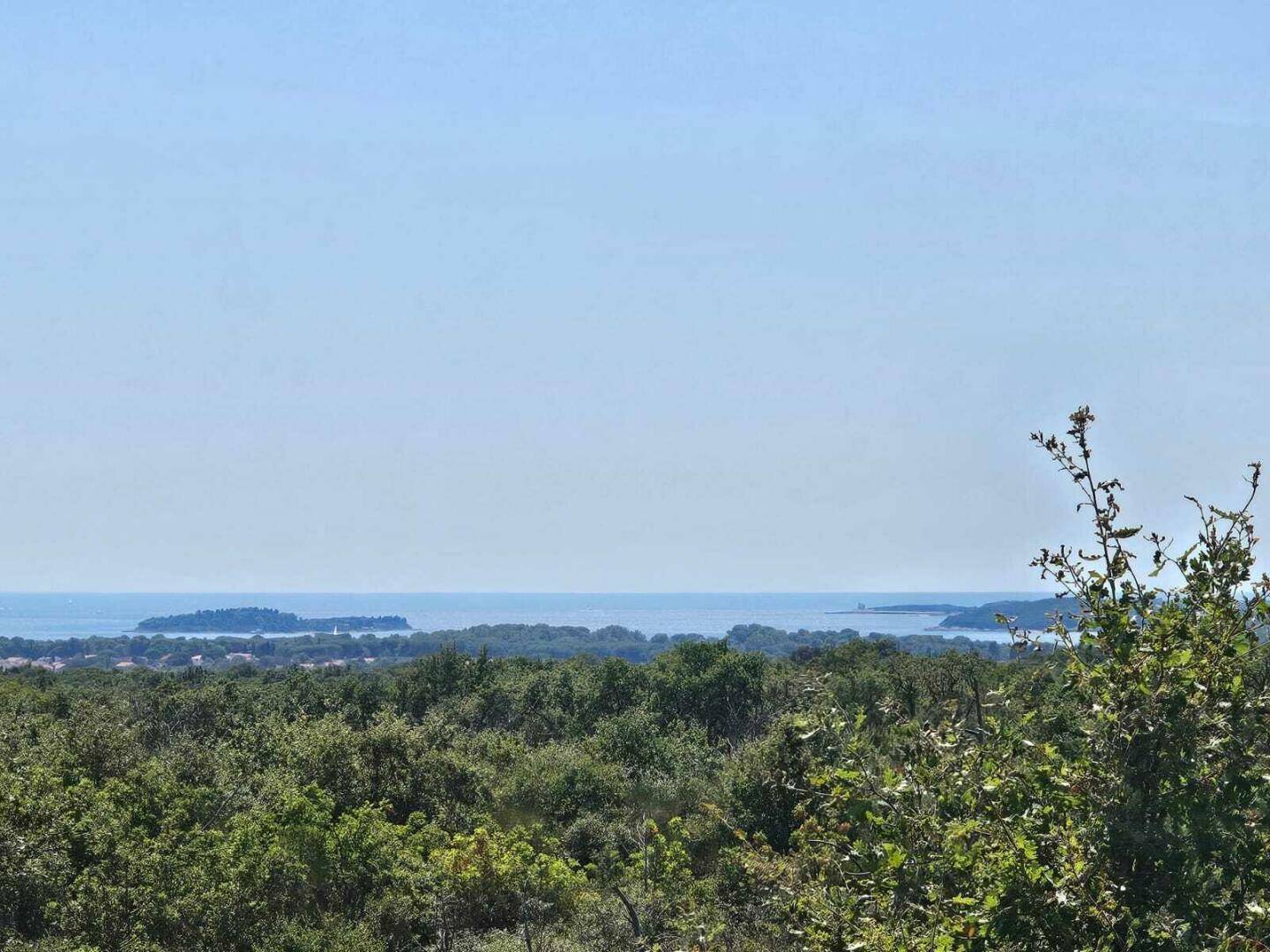 Weitblick über eine grüne Landschaft mit üppiger Vegetation bis zum weiten Meer und entfernten Inseln unter blauem Himmel.